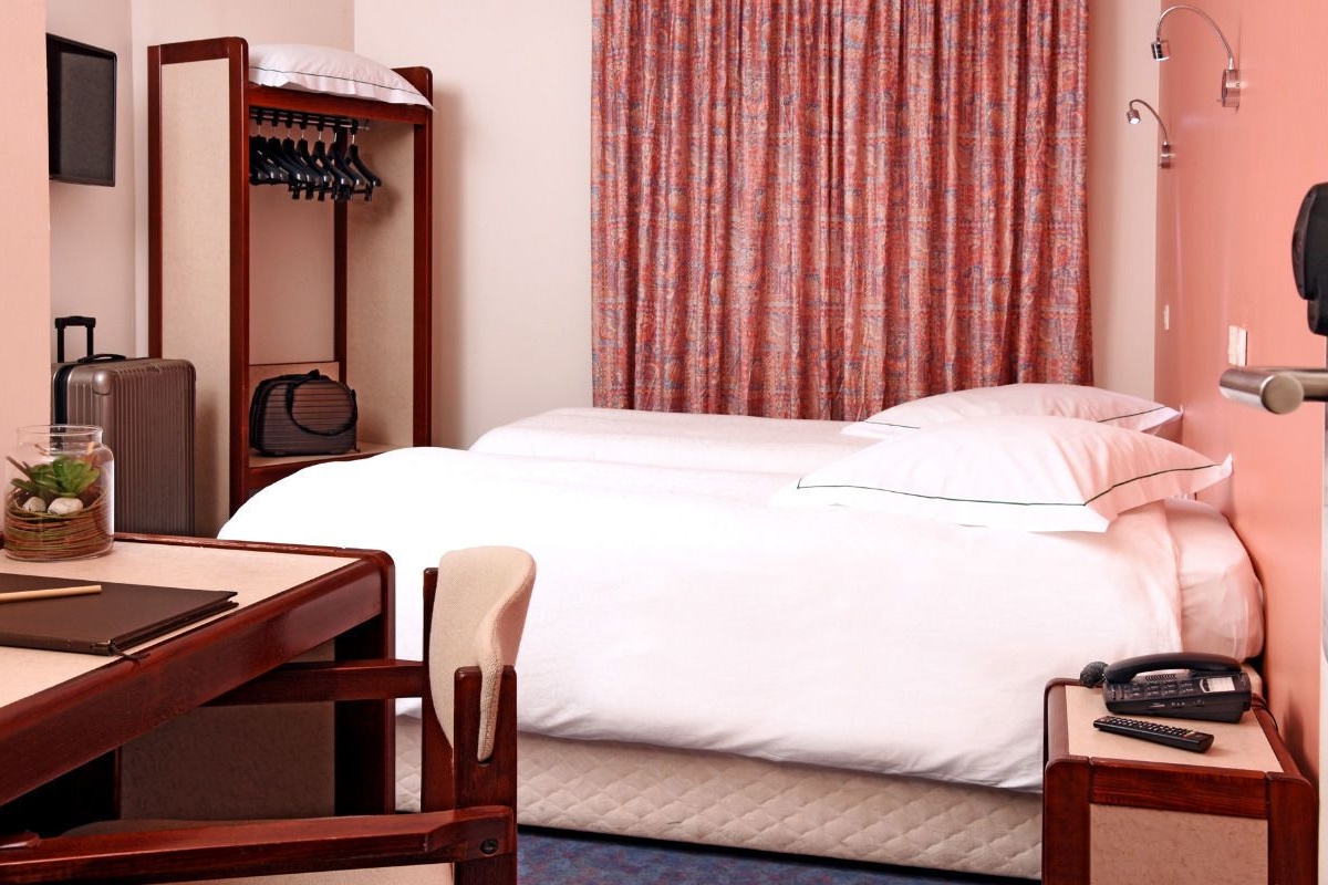 A hotel room with a bed, a desk with a chair, a coat rack, a suitcase, and a remote control on a nightstand.