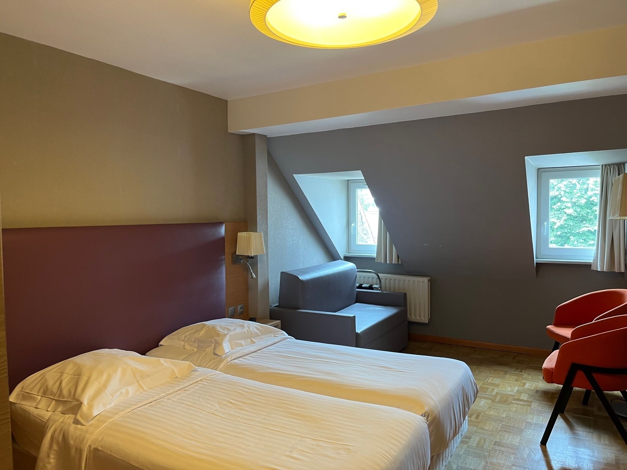 A hotel room with two beds, a desk, a chair, a window, and a lamp.