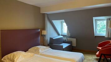 A hotel room with two beds, a desk, a chair, a window, and a lamp.