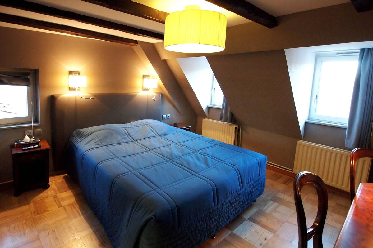 Ein Schlafzimmer mit Bett, Nachttischlampen, Heizkörper, einem Fenster mit Vorhängen, Holzboden und Tisch mit Stuhl.
