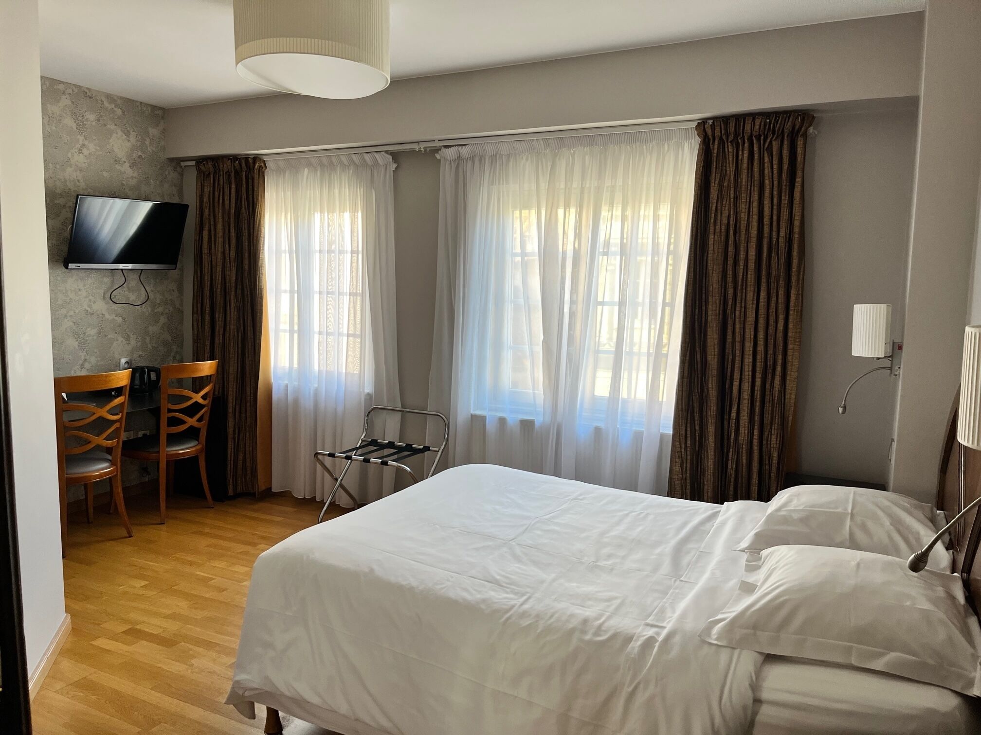 A hotel room with a bed, a TV mounted on the wall, a desk with chairs, and large windows with curtains.