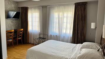 A hotel room with a bed, a TV mounted on the wall, a desk with chairs, and large windows with curtains.