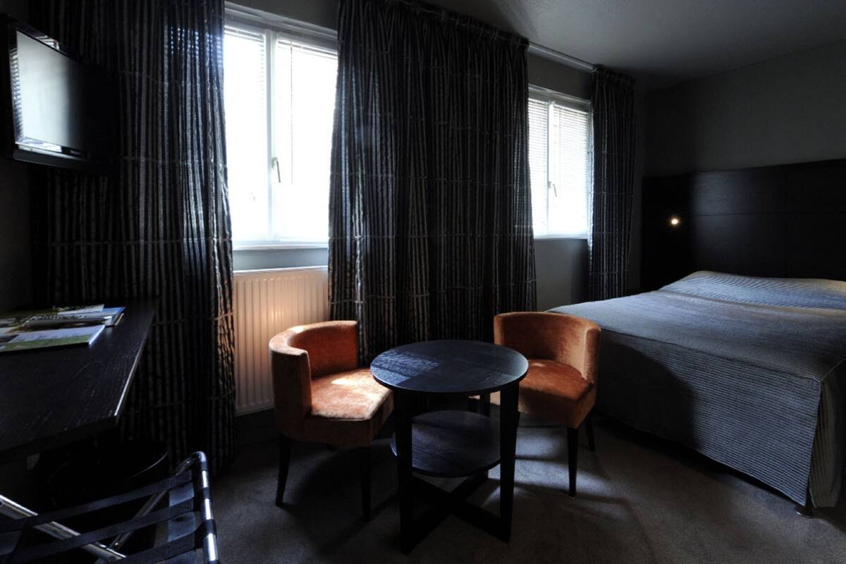 Ein Hotelzimmer mit Bett, zwei Sesseln, einem kleinen Tisch, einem Fernseher und einem Fenster mit Vorhängen.