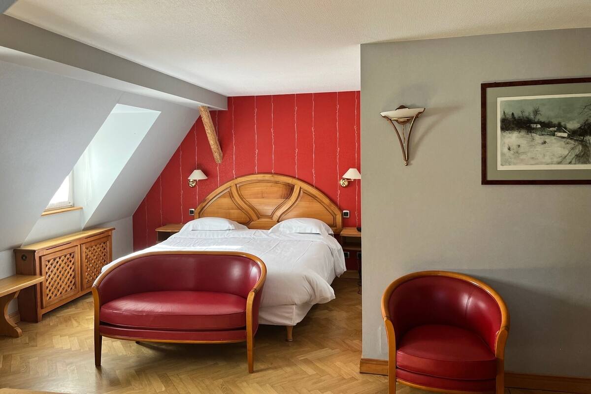 Ein Schlafzimmer mit schrägen Wänden, einem Holzbett, zwei roten Ledersesseln, einem Nachttisch und einer Wandleuchte.