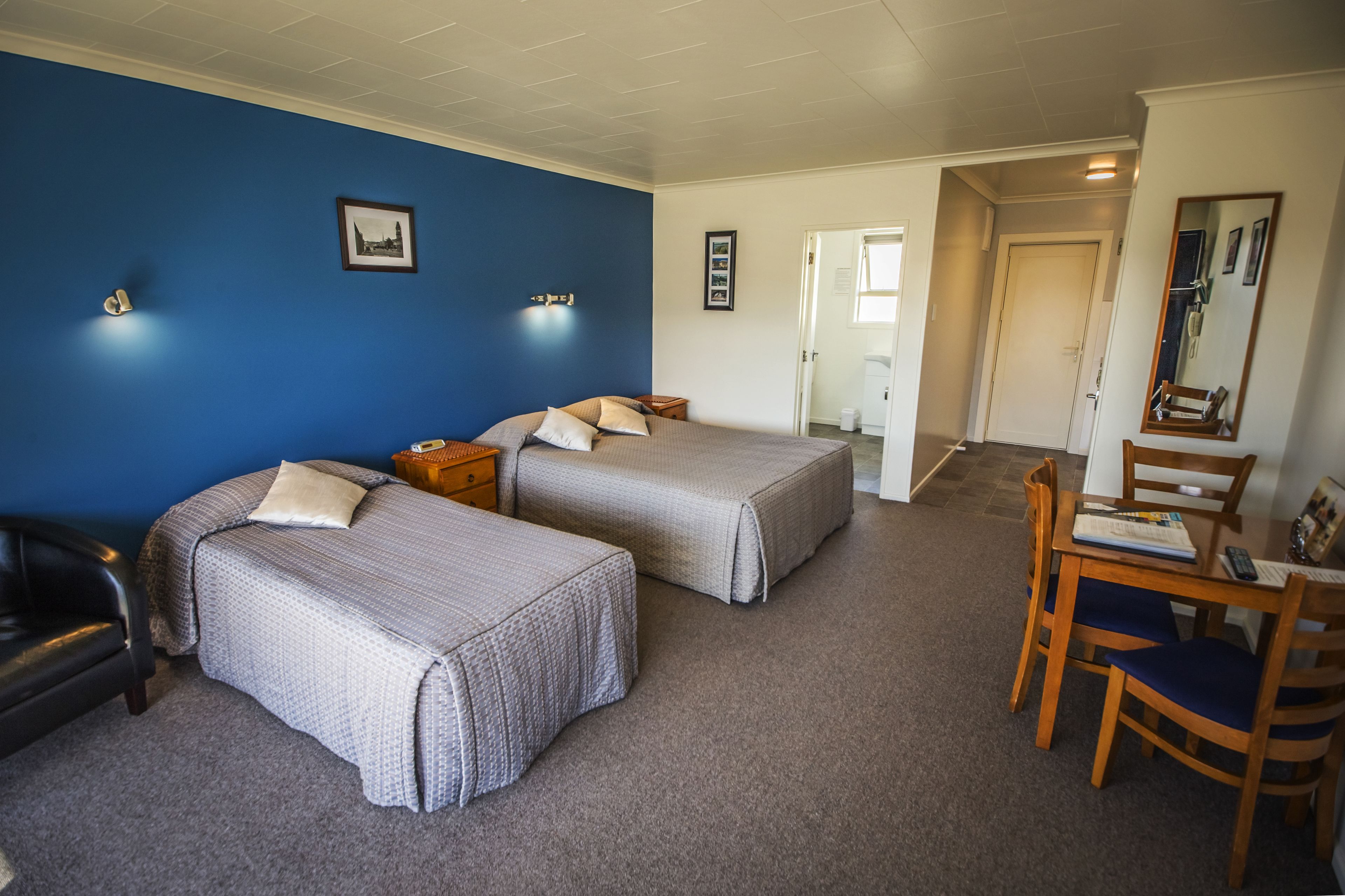 A hotel room with two beds, a desk with a chair, a mirror, and a door leading to another room.