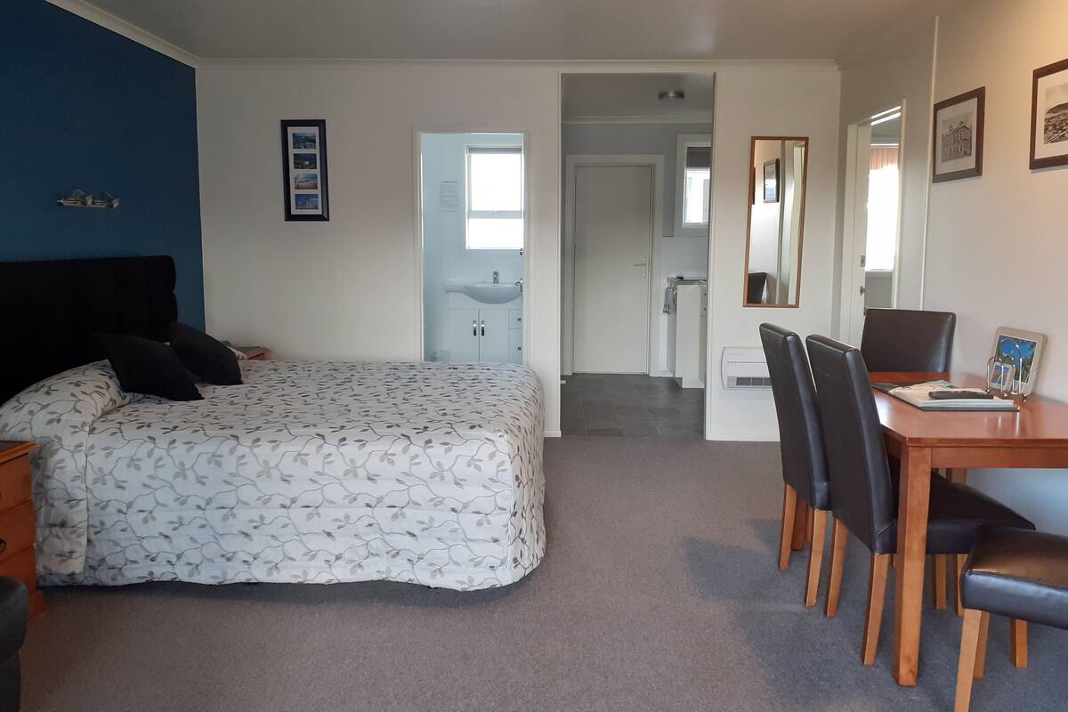 A bedroom with a bed, a desk with chairs, a mirror, and a bathroom visible through an open door.