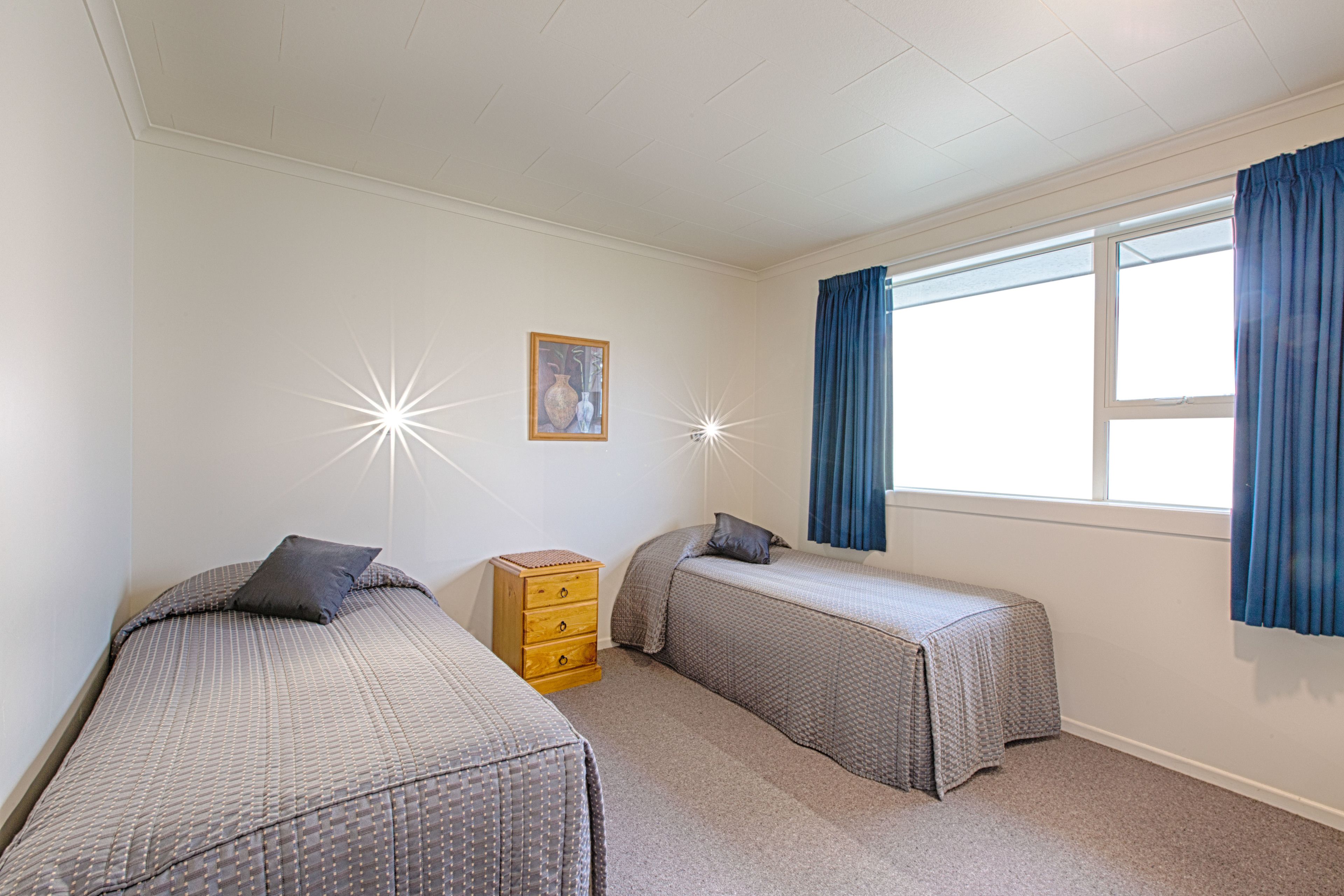 A bedroom with two single beds, a wooden dresser, a framed picture on the wall, and a window with blue curtains.