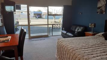 A hotel room with a bed, a desk with a chair, a leather sofa, a nightstand with a lamp, and a view of the street through a glass door.