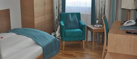 A hotel room with a bed, a teal armchair, a small table, and a desk with a phone and remote control.