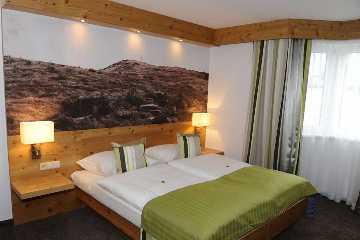 A hotel room with a wooden bed, two bedside lamps, a large wall mural, and a window with curtains.