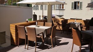 Un patio al aire libre con mobiliario de mimbre y un paraguas grande, con vista a un edificio con balcones y ventanas.