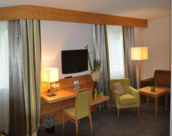 A hotel room with a wooden desk, a television, a small table with a lamp, a green armchair, and a sofa. - Goldener Adler Wattens (Wattens)