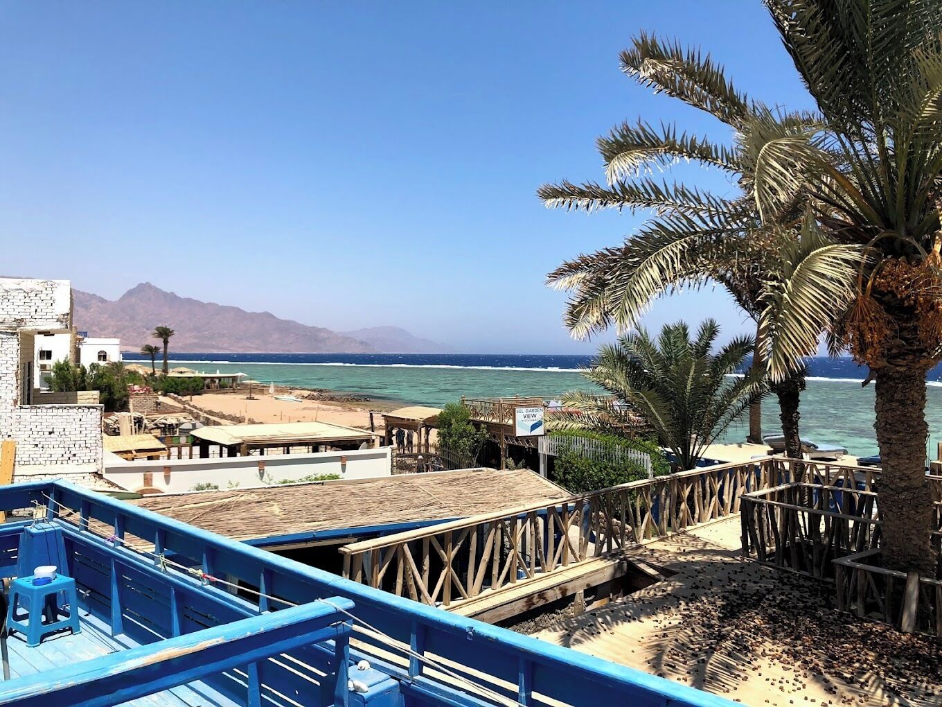 Vista costera con una terraza de madera, palmeras y una playa al fondo.