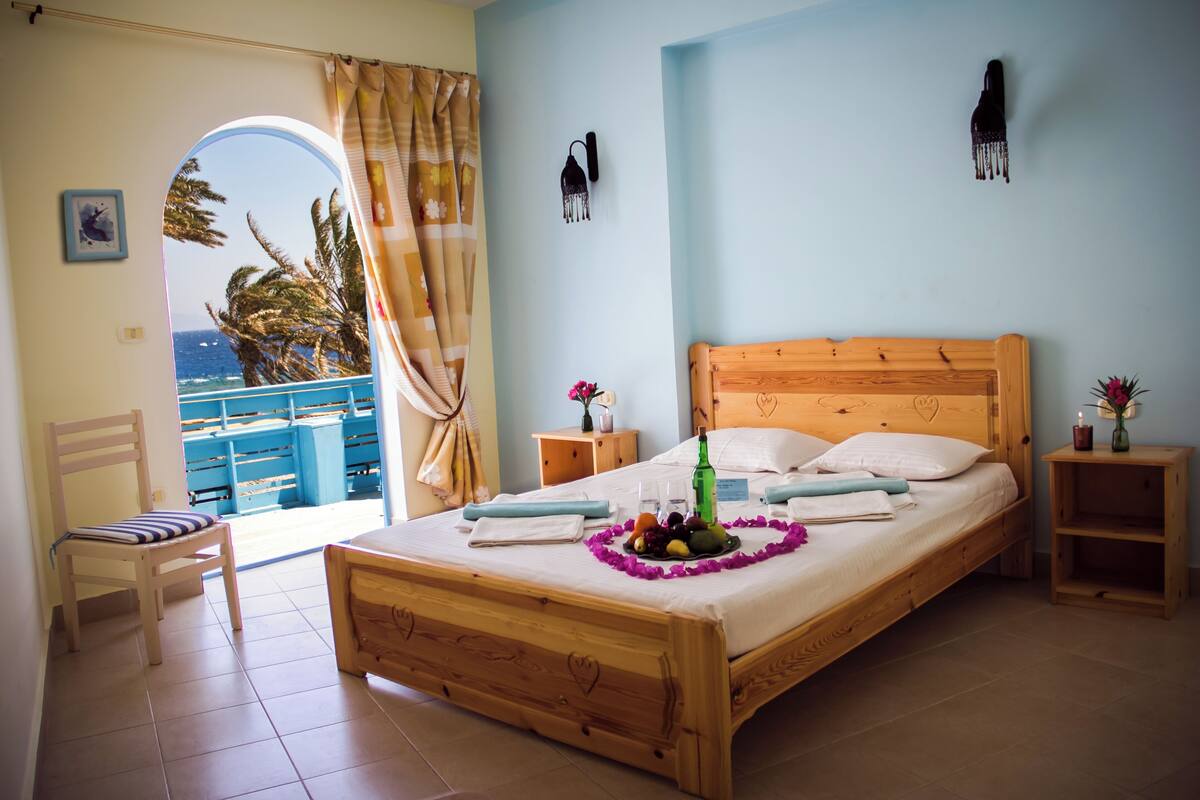 Un dormitorio con cama de madera, vista al mar a través de un arco, una mesita de noche con un florero y un buró con lámpara.