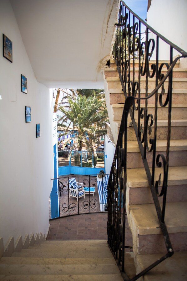 Una escalera con barandillas de metal decoradas lleva a un espacio al aire libre con una mesa y sillas azules, palmeras y vistas a una calle.