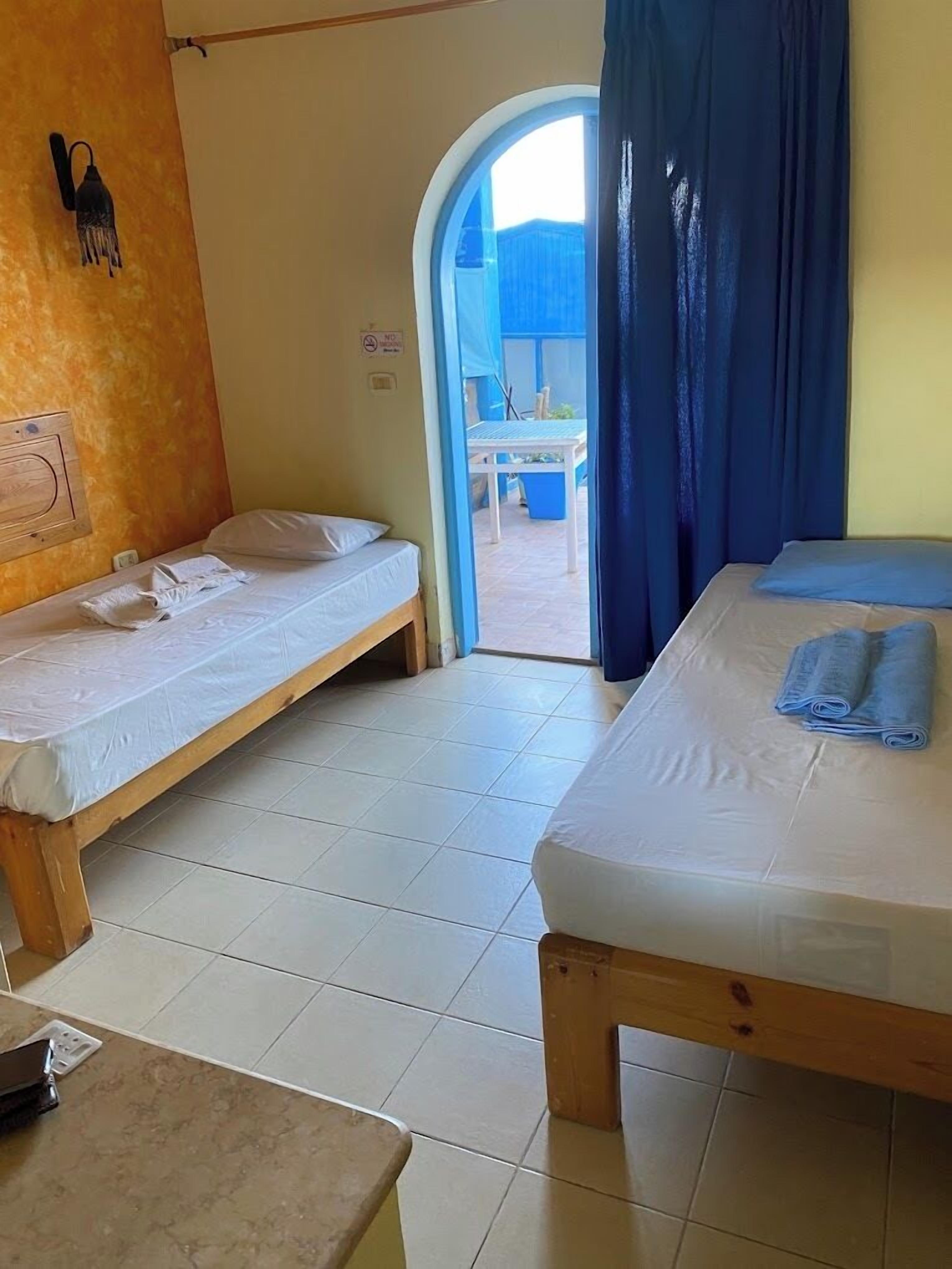 A room with two wooden beds, a small table, and a view of an outdoor area through a doorway.