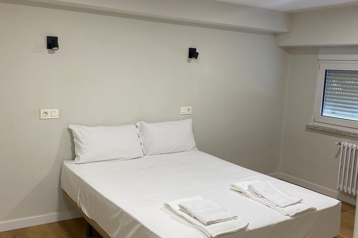 Habitación individual con cama individual, piso de madera, ropa de cama blanca, dos luces empotradas en la pared y ventana con persianas.