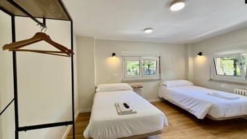 A room with two beds, wooden flooring, a wooden hanger rack, and two windows with views of greenery.
