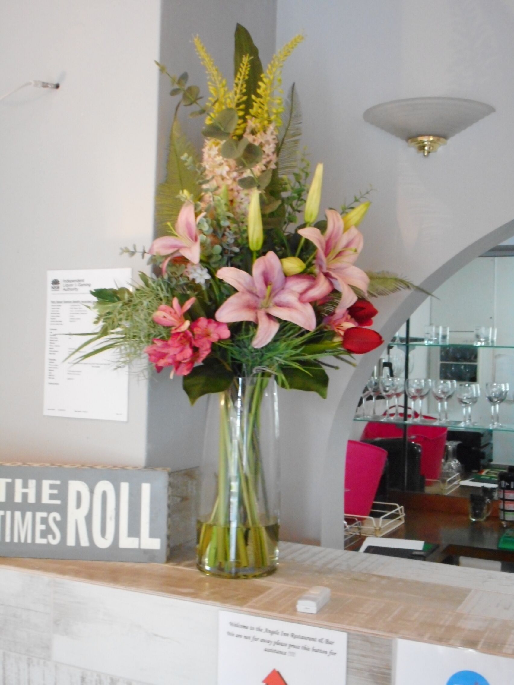 A reception desk with a vase of flowers, a sign reading "THE TIMES ROLL", and a glass display case with wine glasses.