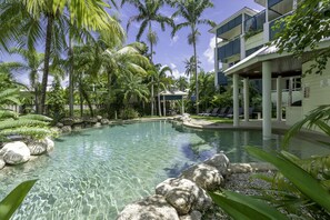 Una piscina cristalina rodeada de rocas y palmeras, con un edificio al fondo que cuenta con balcones y sillones de descanso.