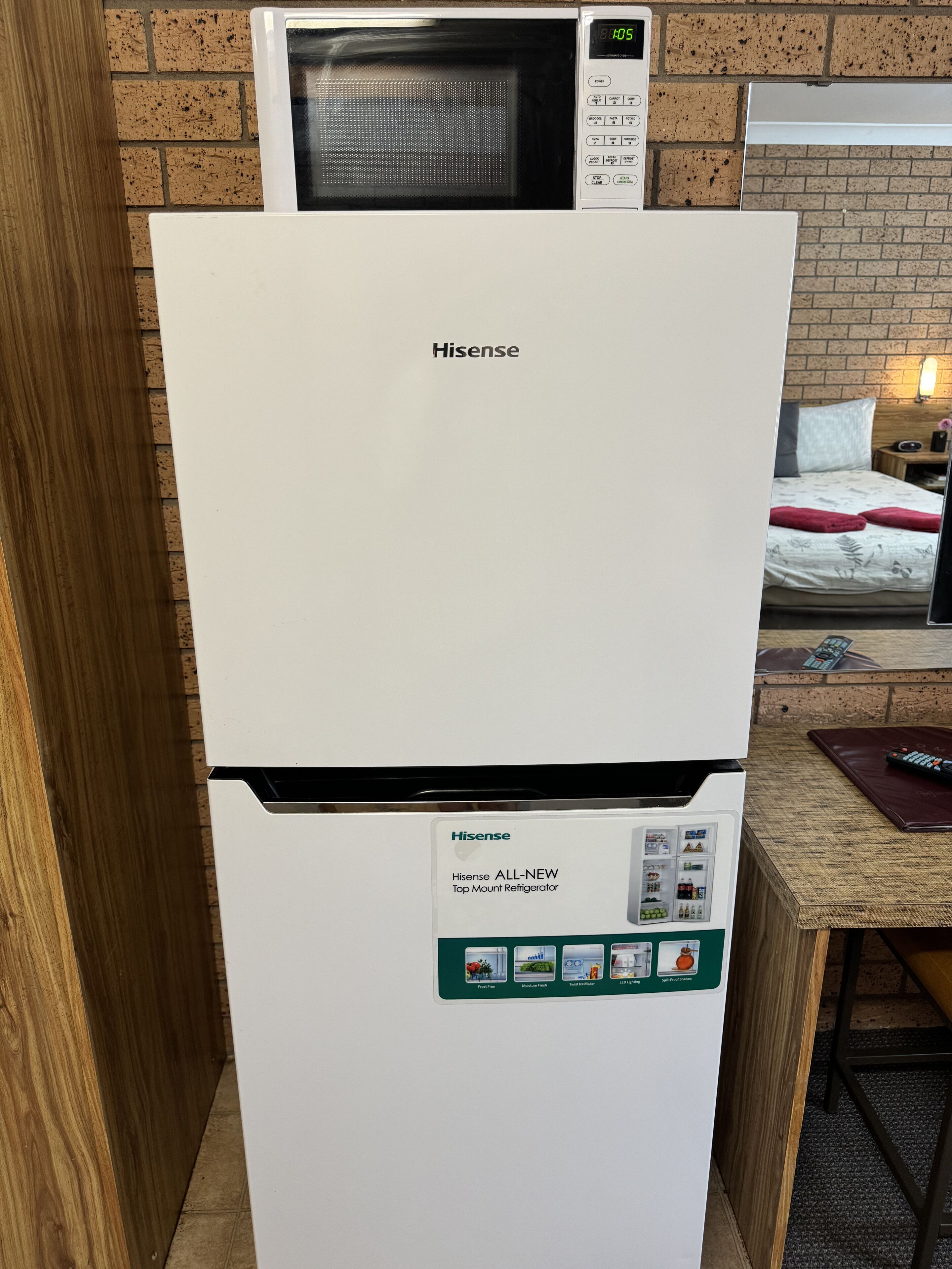Een Hisense inbouwkoelkast in een kamer met een bakstenen muur, een bed met rode kussens en een afstandsbediening op een tafel.