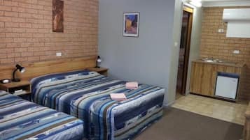 Une chambre avec deux lits, une tête de lit en bois, un petit bureau avec une lampe, un tableau au mur et un coin cuisine.