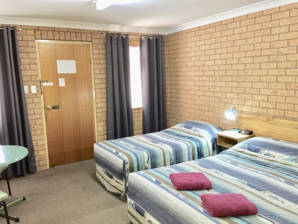 Una habitación con dos camas, una mesita, una lámpara y una pared de ladrillo.