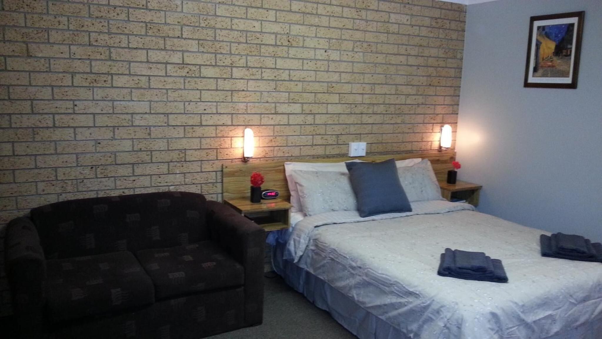 Een hotelkamer met een metselwerk muur, een bed met een witte dekbedovertrek, twee nachtlampjes, een donkere fauteuil en een klein tafeltje met een rode bloem.