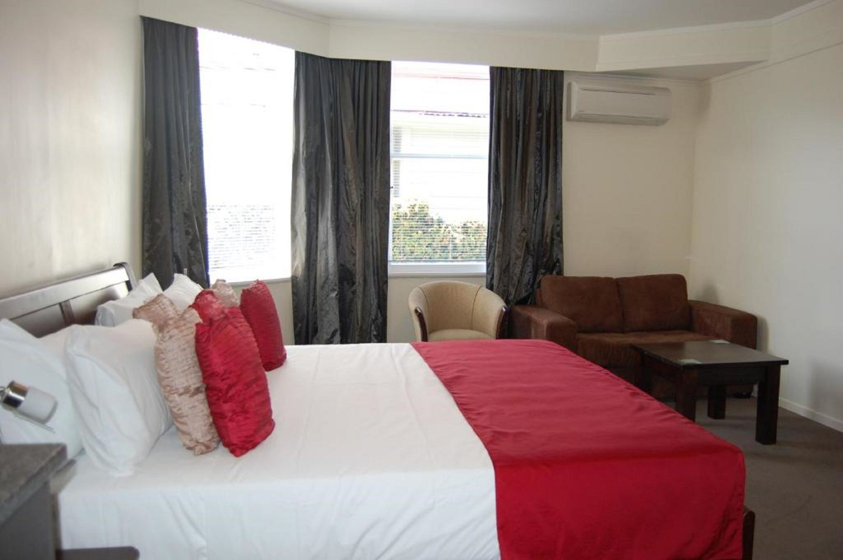 Ein Hotelzimmer mit einem Bett, einer Couch, einem Sessel und einem kleinen Tisch. Ein Fenster mit Vorhängen und eine Klimaanlage an der Wand sind ebenfalls vorhanden.