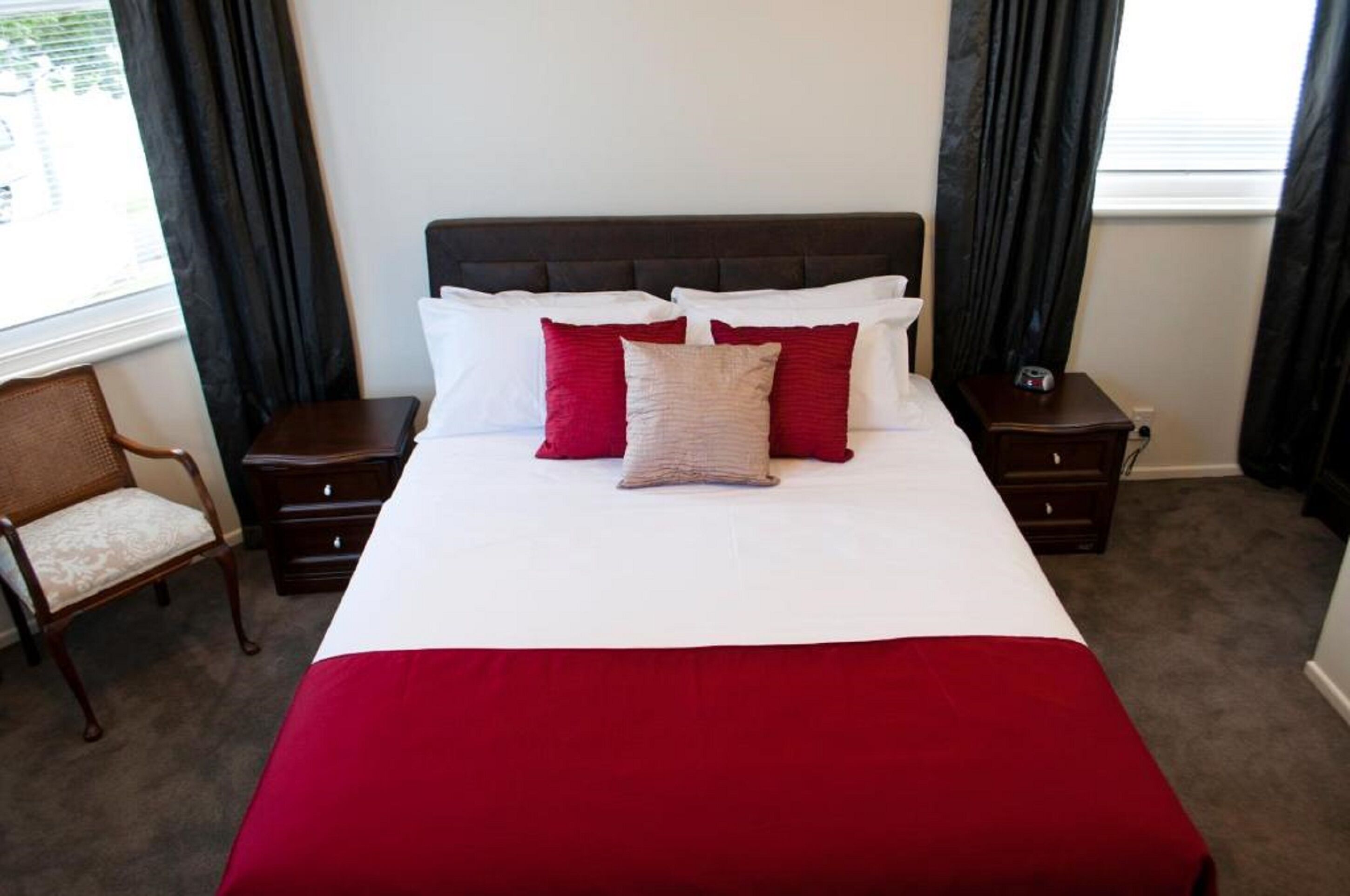 Una cama bien hecha con una colcha roja y blanca, dos mesitas de noche con un reloj y una silla con cojín.