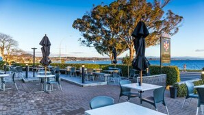 An outdoor cafe with tables and chairs, a sign with a flag, and a view of the sea. - Twin Peaks Lakeside Inn (Taupo)