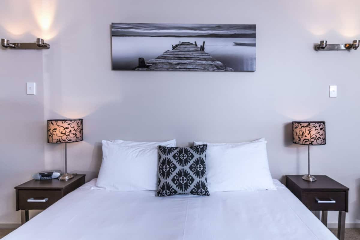A hotel room with a large bed, two bedside tables with lamps, a wall-mounted painting of a pier, and a nightstand with a radio.