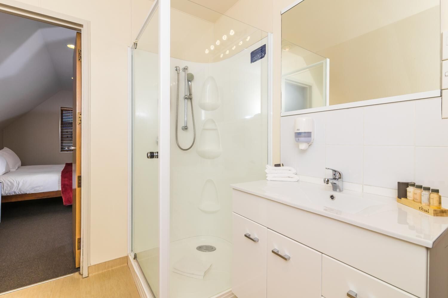 A modern bathroom with a glass-enclosed shower, a white vanity with a mirror, and a bed visible through an open door.