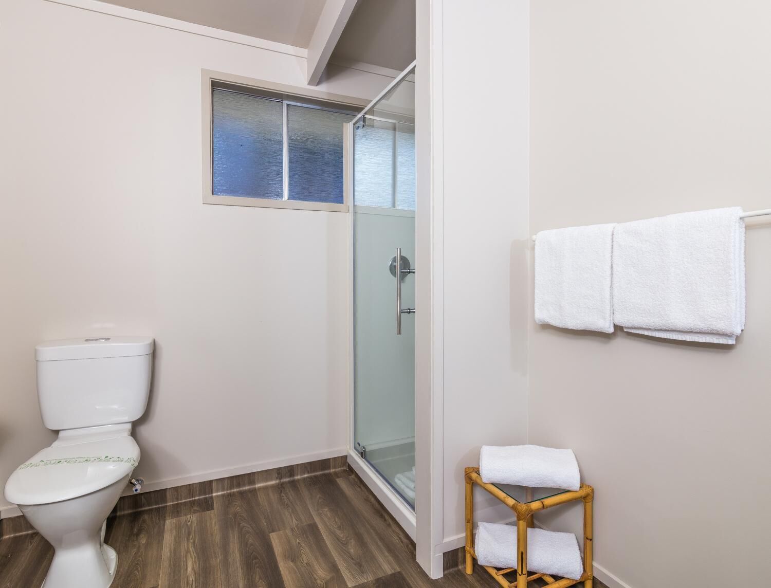 A bathroom with a toilet, a glass shower enclosure, and a small wooden stool with a towel.