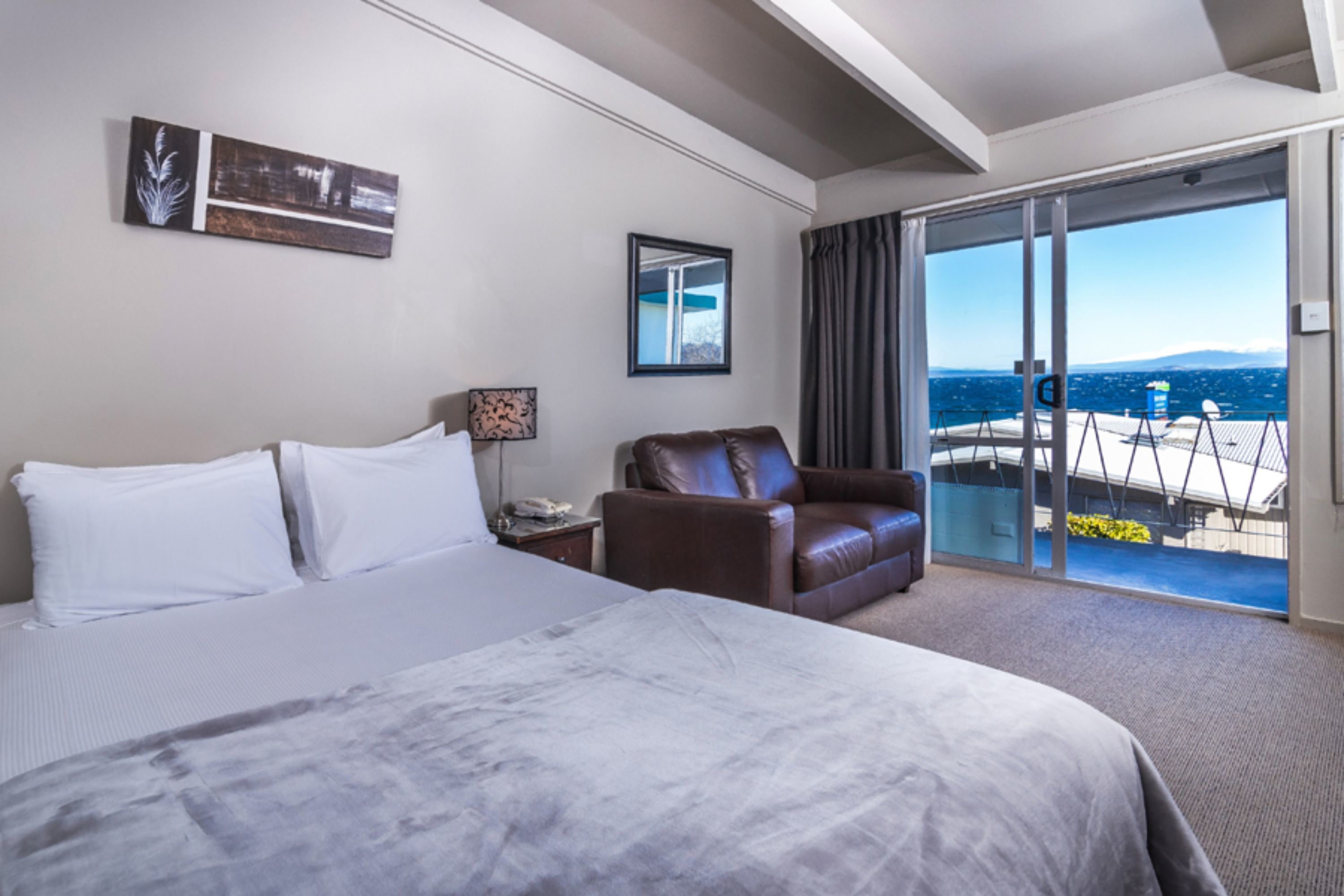 A hotel room with a large bed, a leather armchair, a balcony with a view of the sea, and a sliding glass door.