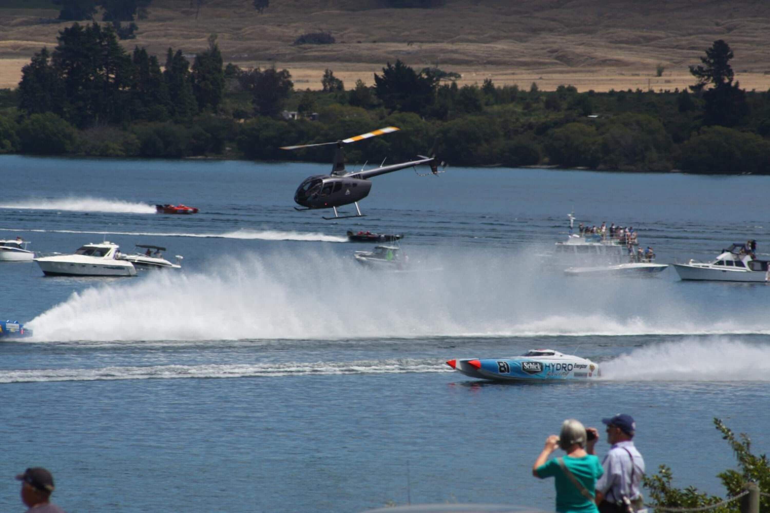 A helicopter flying over a lake with boats and people on the shore.