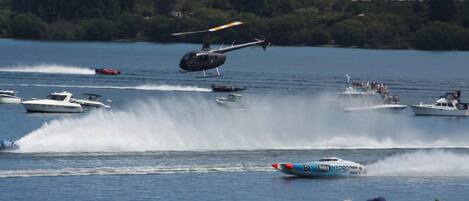 A helicopter flying over a lake with boats and people on the shore.