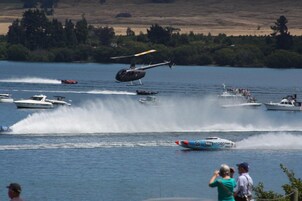 Un helicóptero volando sobre un lago con barcos y gente en la orilla.