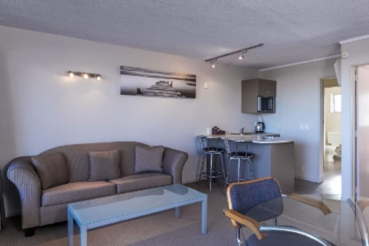 A living room with a sofa, a coffee table, and a chair. There is a kitchen area with a microwave and a bar stool.