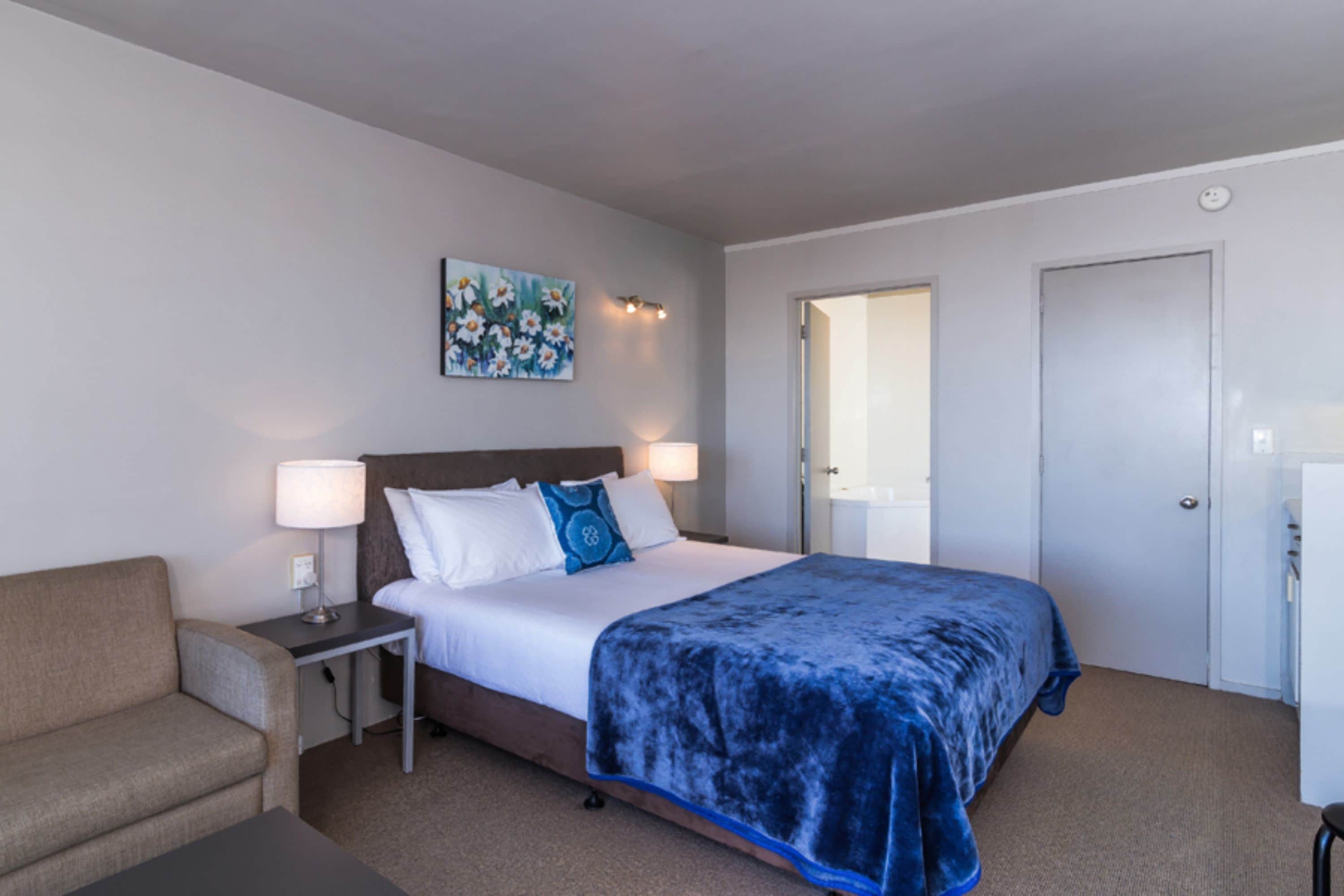 Una habitación de hotel con cama, mesitas de noche, una silla, un cuadro en la pared y una puerta.