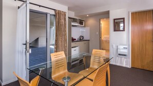 A hotel room with a glass dining table, wooden chairs, a kitchenette, a bathroom, and a sliding door leading to a balcony. - Twin Peaks Lakeside Inn (Taupo)