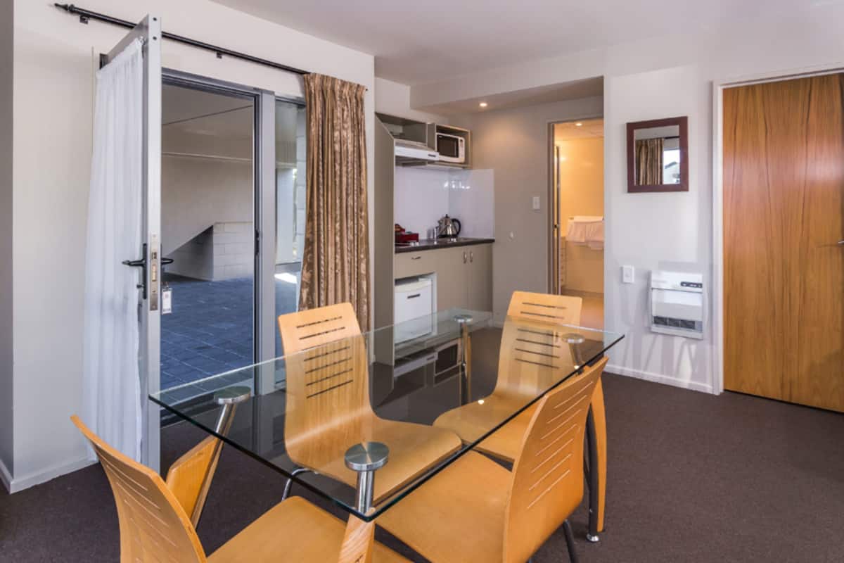 A hotel room with a glass dining table, wooden chairs, a kitchenette, a bathroom, and a sliding door leading to a balcony.