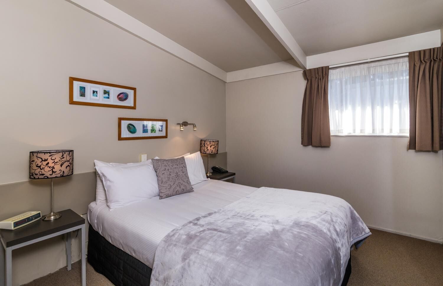 Habitación de hotel con cama, mesitas de noche, lámparas, una ventana con cortinas e imágenes enmarcadas en la pared.