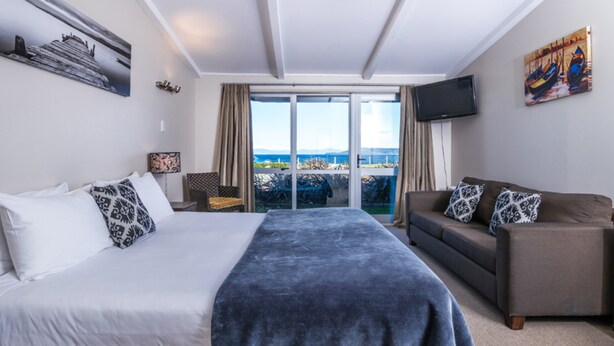 A hotel room with a bed, a sofa, a TV, and a balcony with a view. - Twin Peaks Lakeside Inn (Taupo)
