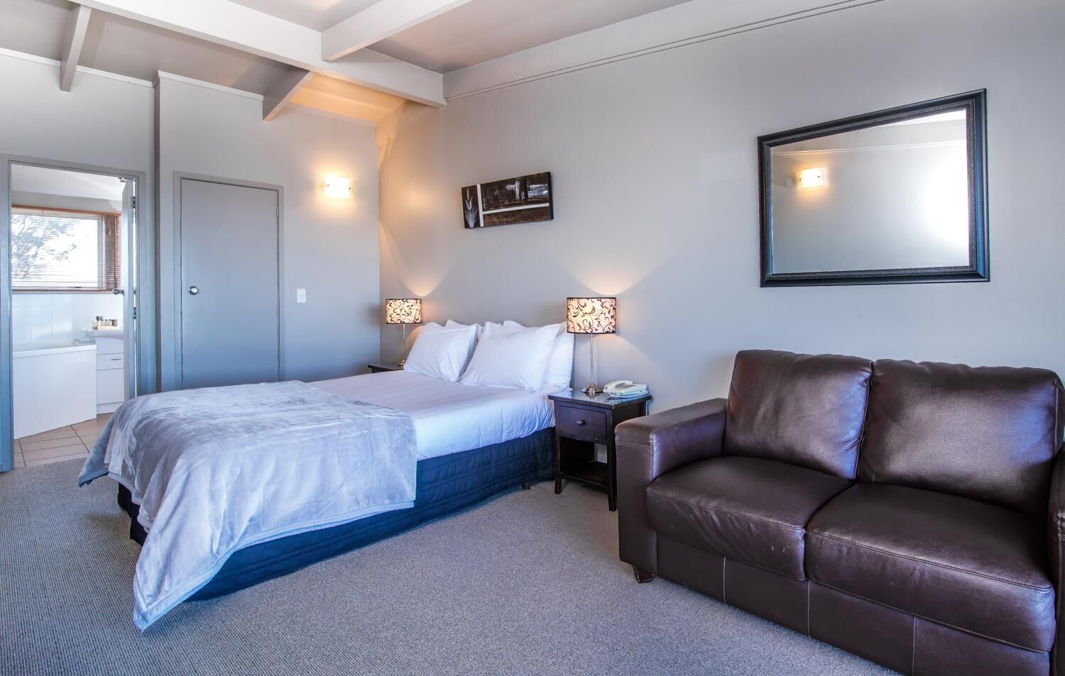 A bedroom with a bed, a brown leather sofa, two bedside tables with lamps, a wall-mounted mirror, and a door leading to another room.