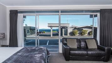 A hotel room with a bed, a leather sofa, and a large sliding glass door leading to a parking area and a building with a view of the sea.