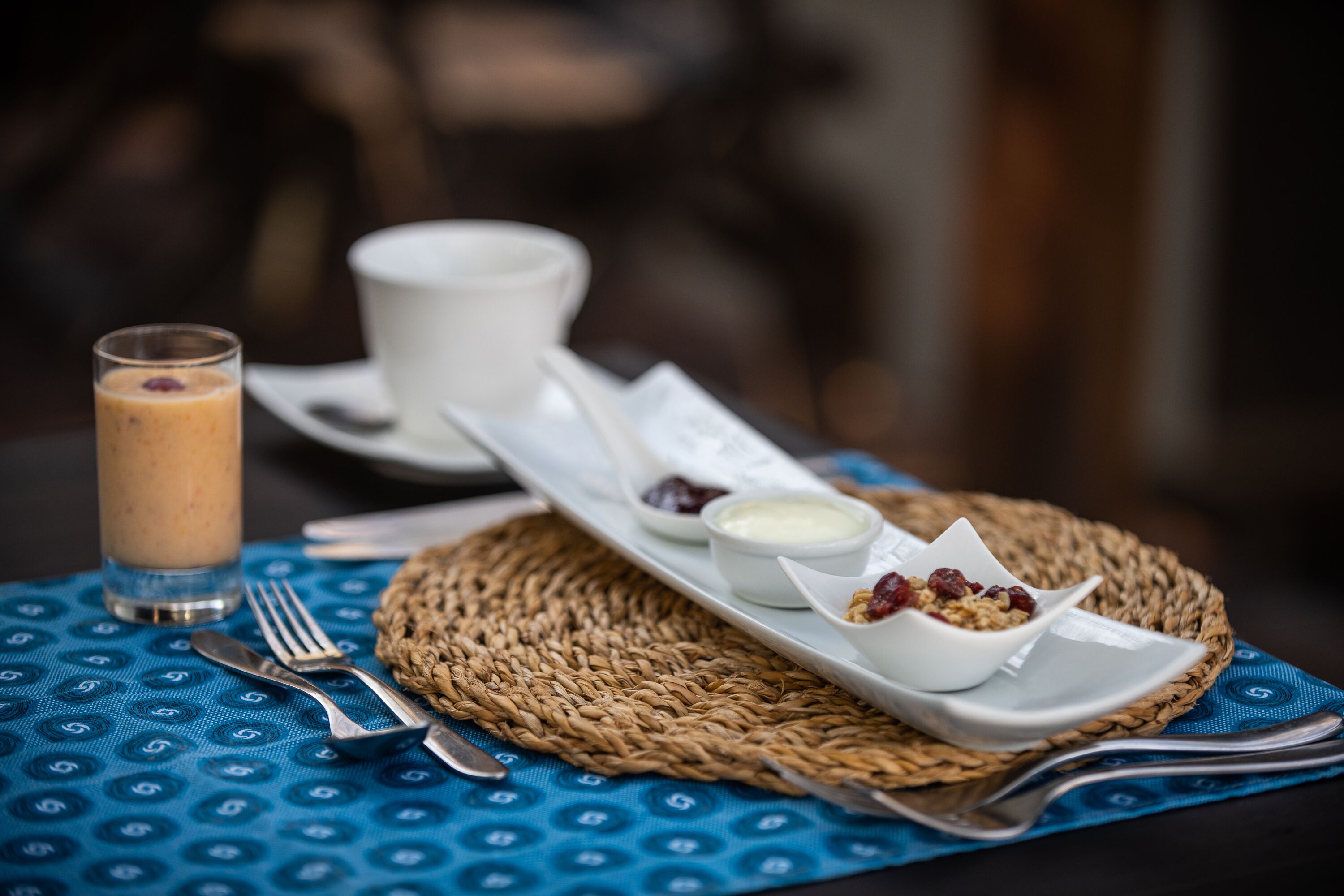 A breakfast setting with a woven placemat, a glass of smoothie, a white cup, and a plate with yogurt and granola.