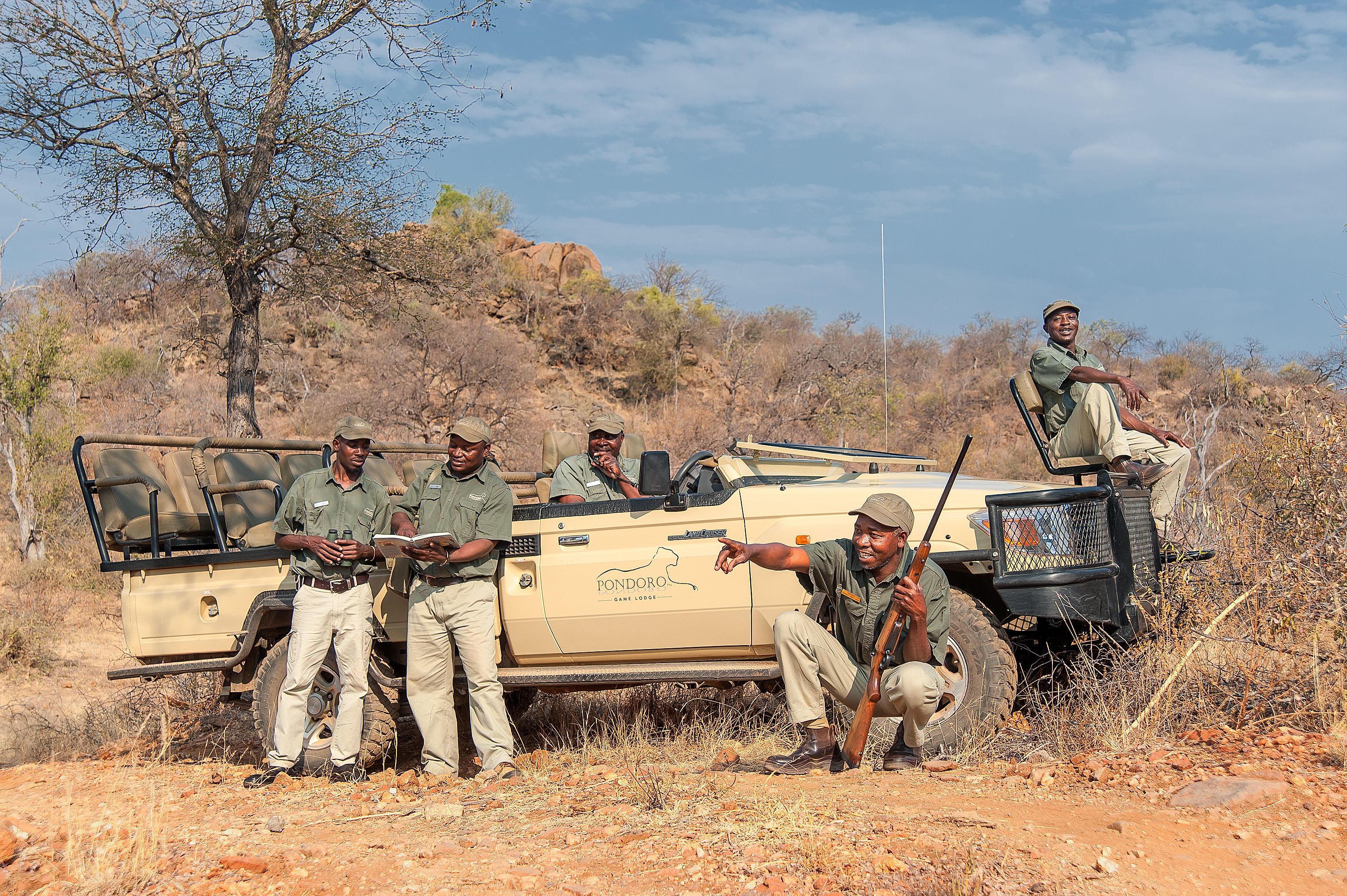 A safari vehicle with four people standing and one sitting, surrounded by dry, rocky terrain and sparse vegetation.
