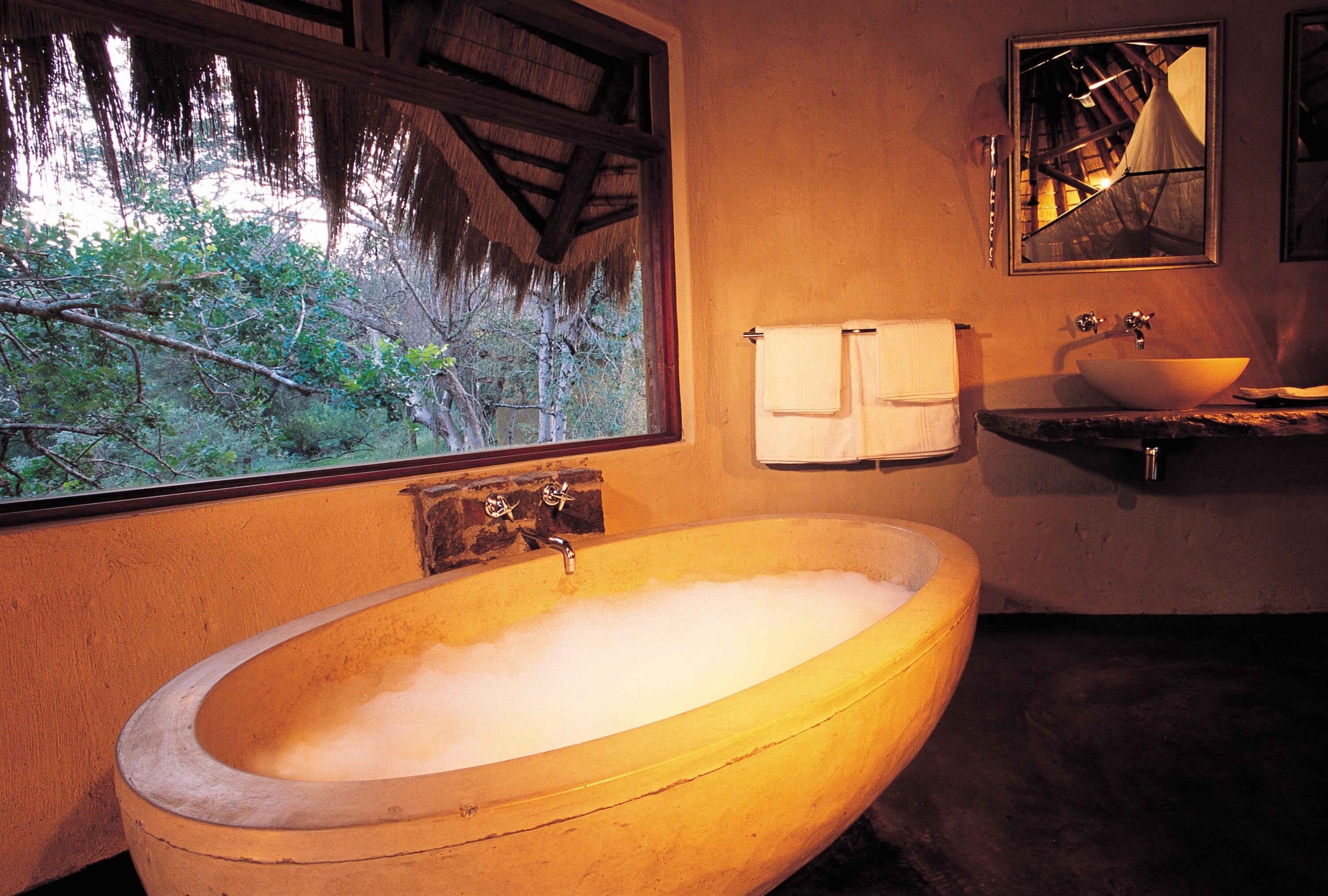 A large oval bathtub with a wooden exterior, a window with a view of trees, a mirror, and a sink with a basin.