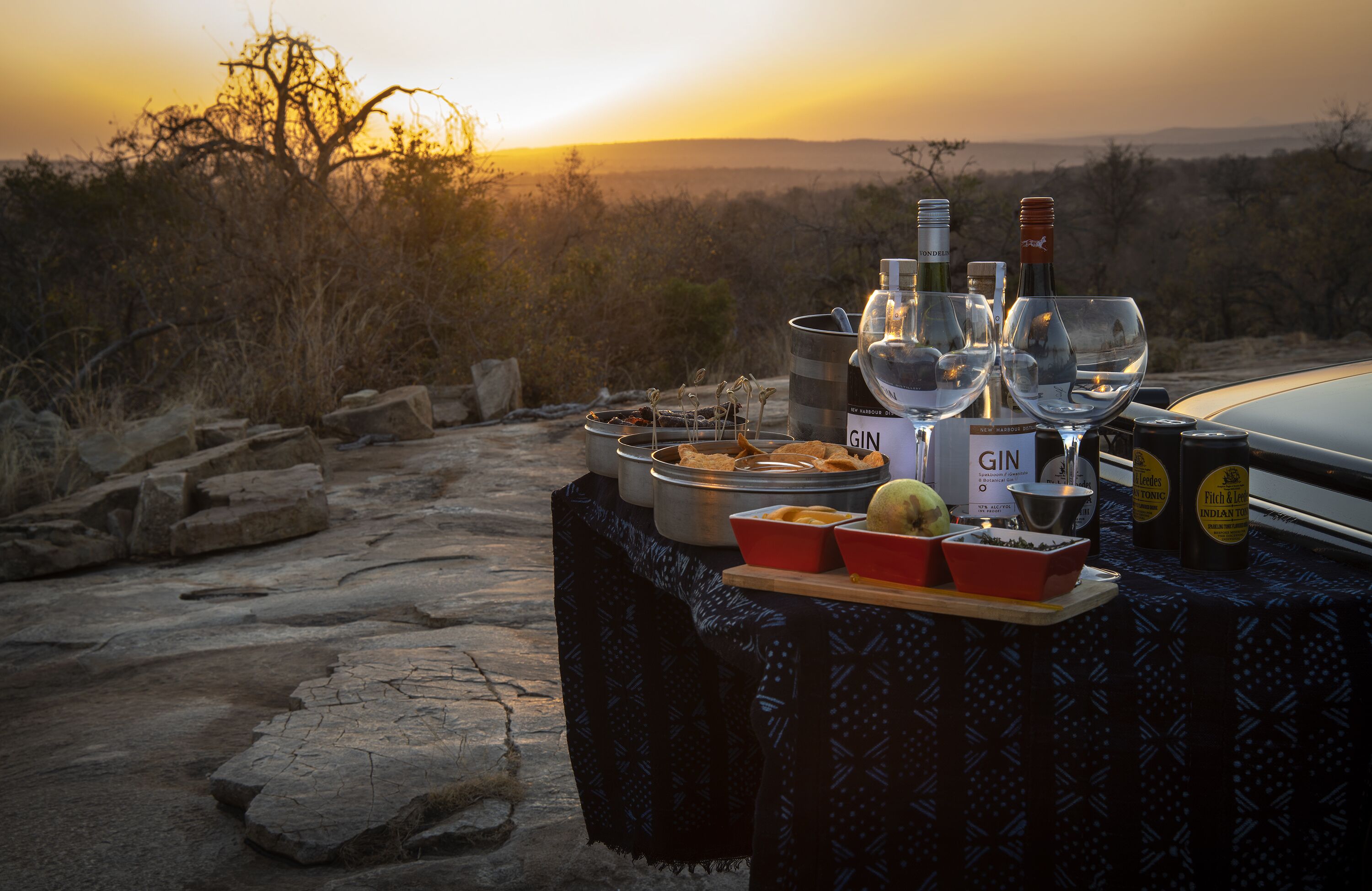A table set outdoors with a sunset view, containing bottles, glasses, and food containers.